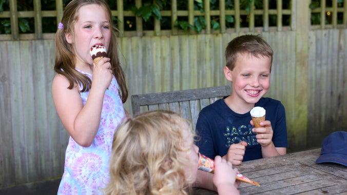 Three children sitting around a table. Two are eating ice creams in a cone and the third has a lolly.